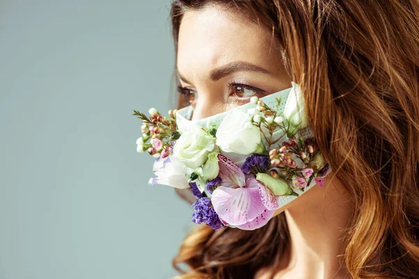 Atractiva y joven mujer en mascarilla con flores aisladas en gris - foto de stock