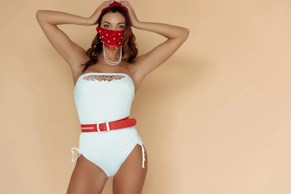 Stylish girl in bathing suit and mask touching hair and standing on beige — Stock Photo