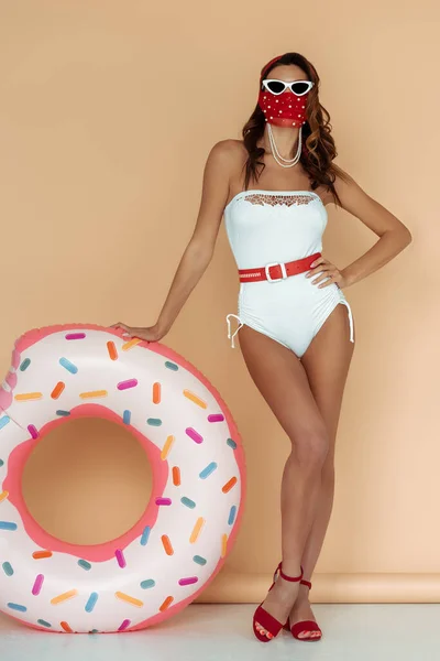 Stylish girl in sunglasses, mask and bathing suit standing with hand on hip near inflatable ring on beige — Stock Photo