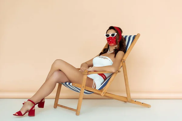 Stylish girl in sunglasses, mask and bathing suit sitting on deck chair on beige — Stock Photo