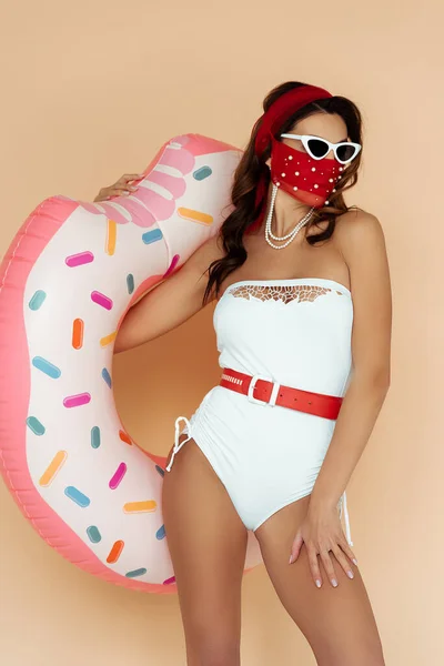 Menina elegante em óculos de sol, máscara e roupa de banho segurando anel inflável isolado no bege — Fotografia de Stock