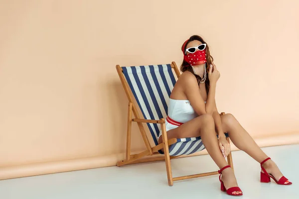 Jeune femme à la mode en lunettes de soleil, masque et maillot de bain assis sur chaise longue sur beige — Photo de stock