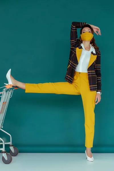 Trendy woman in yellow mask putting leg on empty shopping cart on blue — Stock Photo