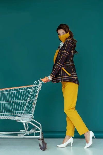 Mujer de moda en máscara amarilla caminando con el carrito de compras vacío en azul - foto de stock