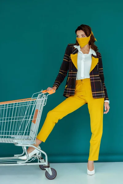 Stylish girl in yellow mask putting leg on empty shopping cart on blue — Stock Photo