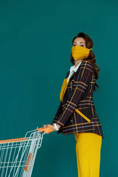 Trendfrau in gelber Maske steht mit leerem Einkaufswagen isoliert auf blau — Stockfoto