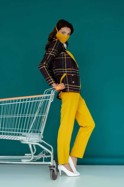Donna alla moda in maschera gialla posa vicino vuoto carrello della spesa su blu — Foto stock
