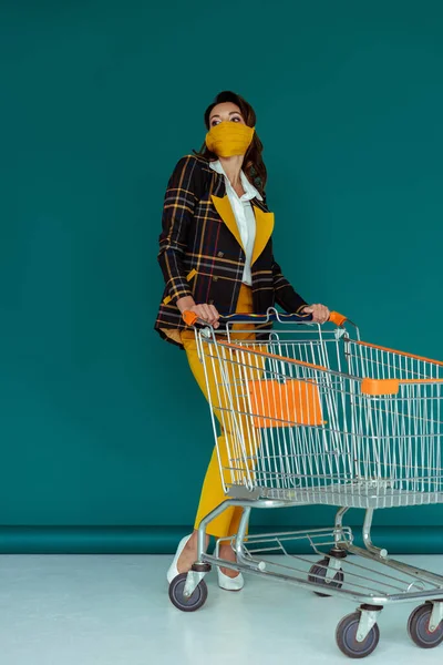 Mujer de moda en máscara amarilla de pie cerca del carrito de compras vacío en azul - foto de stock