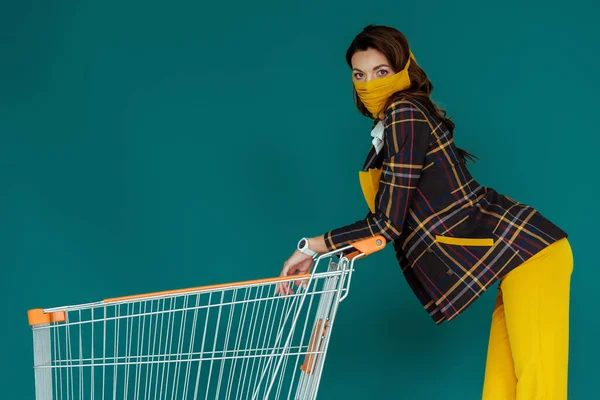 Stylish woman in yellow mask standing with empty shopping cart on blue — Stock Photo