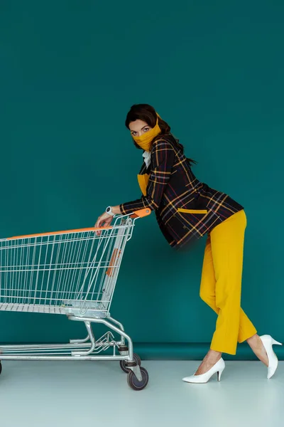 Mulher elegante em máscara amarela e blazer xadrez de pé com carrinho de compras vazio no azul — Fotografia de Stock