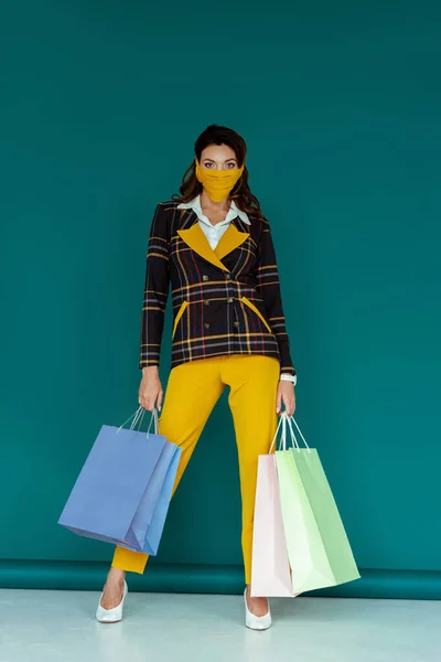 Mujer con estilo en máscara amarilla y chaqueta a cuadros de pie con bolsas de compras en azul - foto de stock