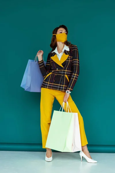 Mujer con estilo en máscara amarilla y chaqueta a cuadros posando con bolsas de compras en azul - foto de stock