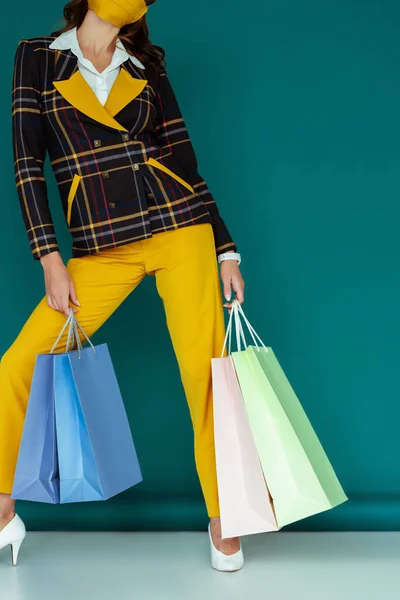 Ausgeschnittene Ansicht einer stilvollen Frau in gelber Maske und kariertem Blazer, die mit Einkaufstaschen auf blauem Grund posiert — Stockfoto