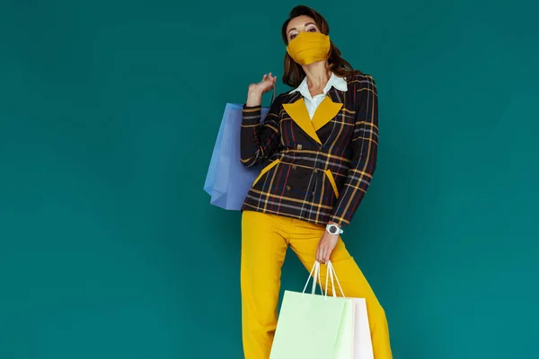 Jeune femme en masque jaune et blazer à carreaux posant avec des sacs à provisions sur bleu — Photo de stock