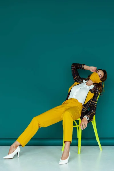 Mujer con estilo en máscara amarilla y chaqueta a cuadros sentado en la silla en azul - foto de stock