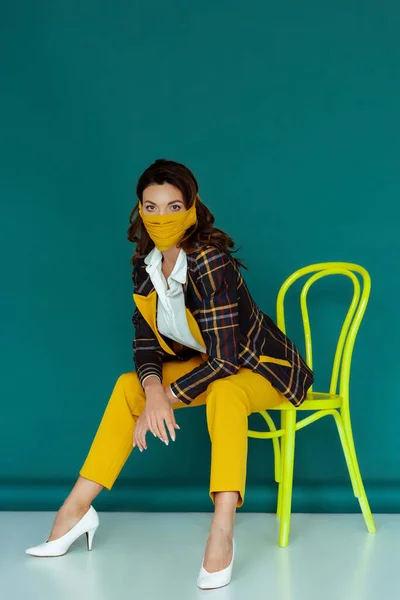 Стильна жінка в жовтій масці і плетений блейзер позує, сидячи на стільці на синьому — стокове фото