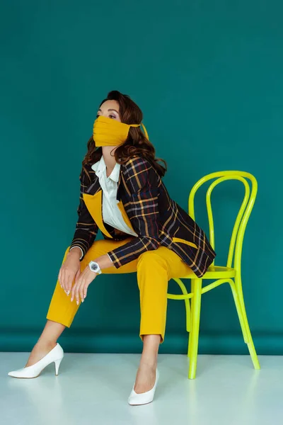 Femme à la mode en masque jaune et blazer à carreaux posant tout en étant assis sur une chaise sur bleu — Photo de stock