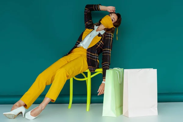 Mujer de moda en máscara amarilla y chaqueta a cuadros posando mientras está sentado en la silla cerca de bolsas de compras en azul - foto de stock