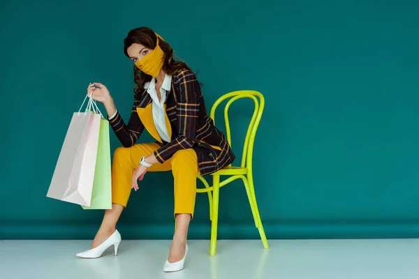 Jeune femme en masque jaune et blazer à carreaux posant tout en étant assis sur une chaise avec des sacs à provisions sur bleu — Photo de stock