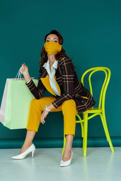 Femme à la mode en masque jaune et blazer à carreaux posant tout en étant assis sur une chaise avec des sacs à provisions sur bleu — Photo de stock