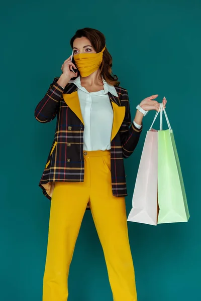 Donna alla moda in maschera gialla e blazer scozzese in posa con shopping bag e parlando su smartphone isolato su blu — Foto stock