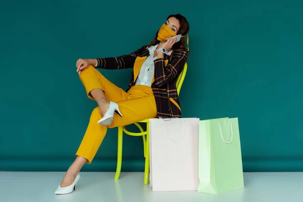 Trendy woman in yellow mask and plaid blazer talking on smartphone and sitting on chair near shopping bags on blue — Stock Photo