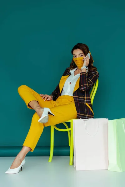 Стильная женщина в желтой маске и клетчатом пиджаке разговаривает на смартфоне и сидит на стуле рядом с магазинами сумки на синий — стоковое фото