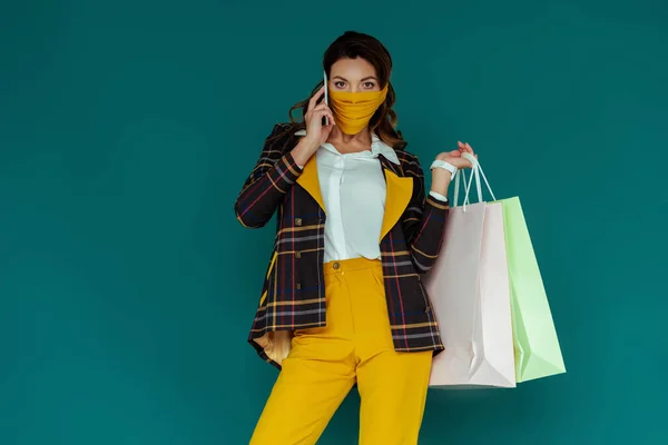 Femme élégante en masque jaune et blazer à carreaux posant avec des sacs à provisions et parlant sur smartphone isolé sur bleu — Photo de stock