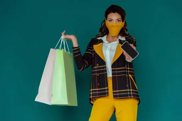 Stylische Frau in gelber Maske und kariertem Blazer mit Einkaufstaschen und Smartphone mit leerem Bildschirm auf blauem Hintergrund — Stockfoto