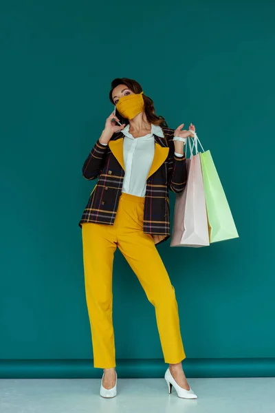Стильная женщина в маске и клетчатом пиджаке позирует с сумками для покупок и разговаривает на смартфоне на голубом — стоковое фото
