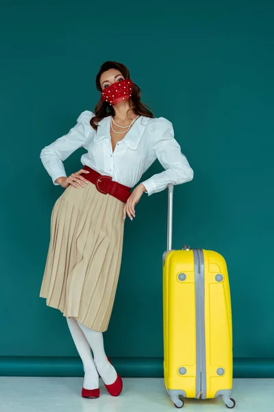 Elegante giovane donna in maschera rossa in piedi con mano sul fianco vicino borsa da viaggio su blu — Foto stock