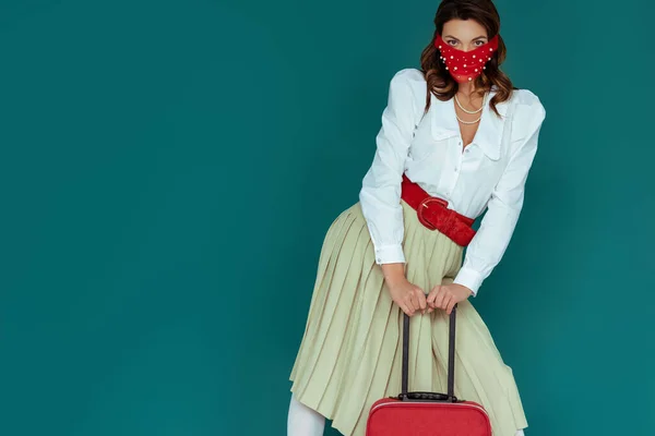 Стильна дівчина в червоній масці, що стоїть біля дорожньої сумки ізольована на синьому — стокове фото