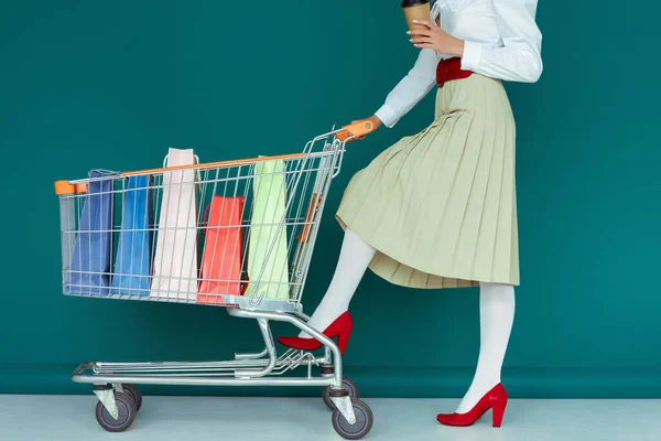 Vista ritagliata della ragazza alla moda che tiene il caffè per andare e in piedi vicino al carrello della spesa con borse della spesa su blu — Foto stock