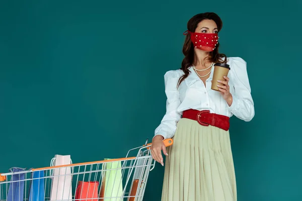 Femme à la mode en masque rouge tenant café pour aller et debout près du panier avec des sacs isolés sur bleu — Photo de stock