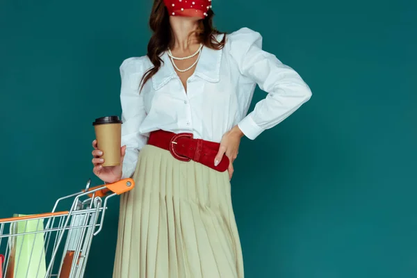 Vista recortada de la chica de moda en máscara roja sosteniendo café para ir y de pie con la mano en la cadera cerca del carrito de la compra con bolsas aisladas en azul - foto de stock