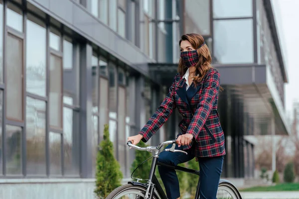 Businesswoman in plaid mask riding bicycle outside — Stock Photo