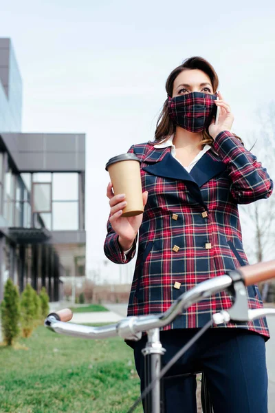 Empresária em xadrez máscara segurando café para ir perto de bicicleta — Fotografia de Stock