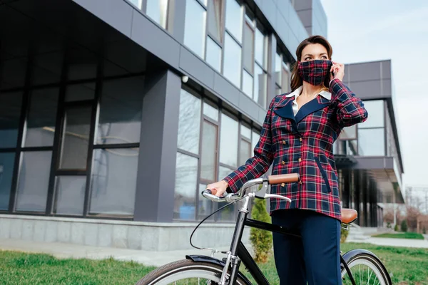 Femme d'affaires en masque à carreaux debout près du vélo à l'extérieur — Photo de stock