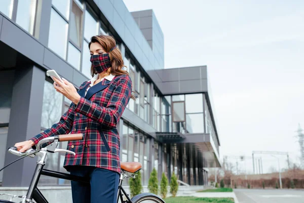 Businesswoman in plaid mask using smartphone near bicycle outside — Stock Photo