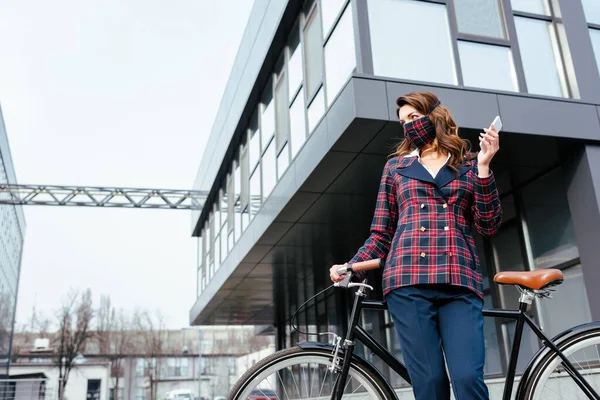 Mujer de negocios en la máscara de cuadros de pie cerca de la bicicleta con teléfono inteligente - foto de stock