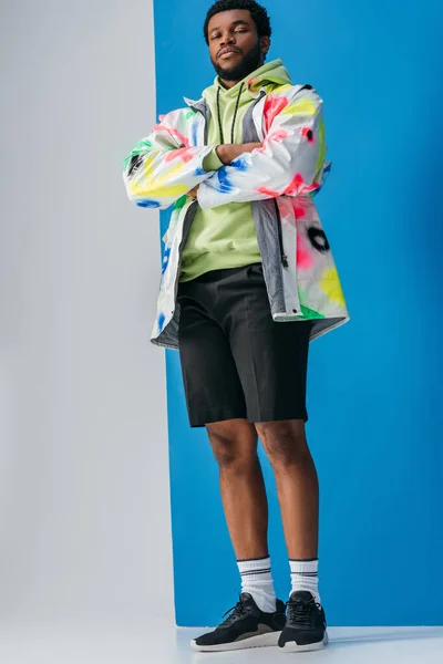 Homme afro-américain à la mode posant avec les bras croisés dans un look futuriste coloré sur gris et bleu — Photo de stock