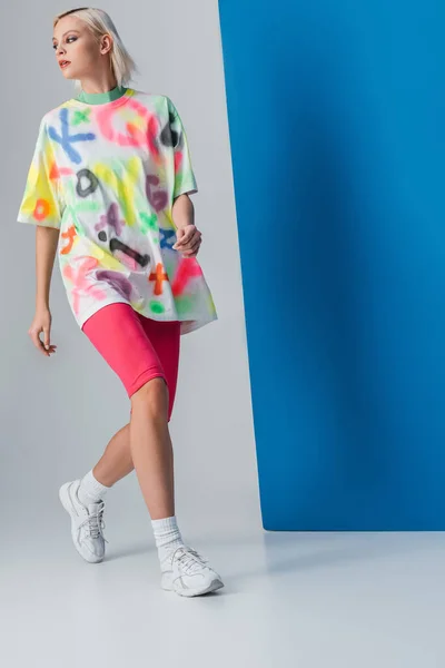 Fashionable woman posing in neon pink bike shorts and colorful t-shirt on grey and blue — Stock Photo