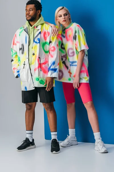 Coppia multietnica alla moda in posa in look futuristico colorato su grigio e blu — Foto stock