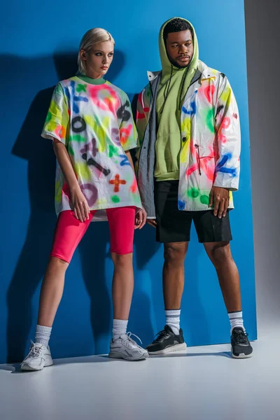 Couple multiculturel élégant posant dans un look futuriste coloré sur gris et bleu — Photo de stock