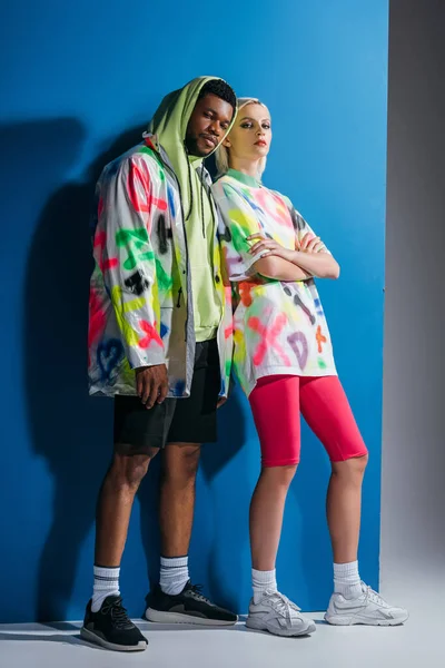 Pareja multicultural de moda posando en mirada futurista colorido en gris y azul - foto de stock