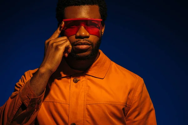 Modelo masculino afroamericano en look futurista y gafas de sol posando sobre azul en luz roja - foto de stock
