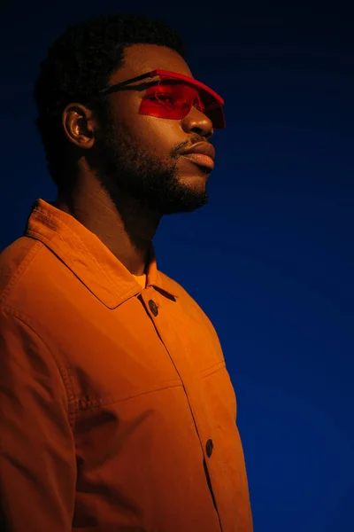 Modisches afrikanisch-amerikanisches Männermodel in futuristischem Look und Sonnenbrille posiert auf blauem Grund im Rotlicht — Stockfoto