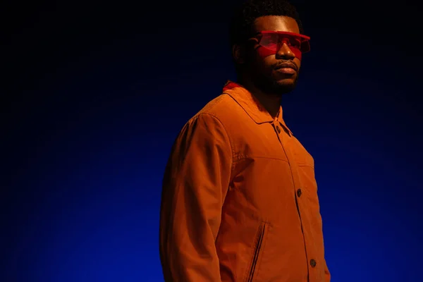 Hombre afroamericano de moda en look futurista y gafas de sol posando sobre azul en luz roja — Stock Photo