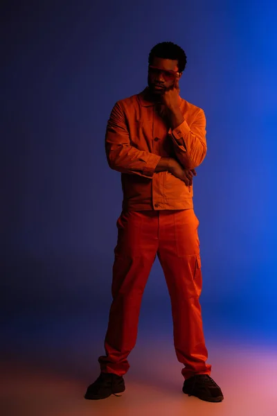 Élégant homme afro-américain au look futuriste et lunettes de soleil posant sur le bleu dans la lumière rouge — Photo de stock