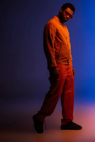 Jeune homme afro-américain au look futuriste et lunettes de soleil posant sur le bleu dans la lumière rouge — Photo de stock